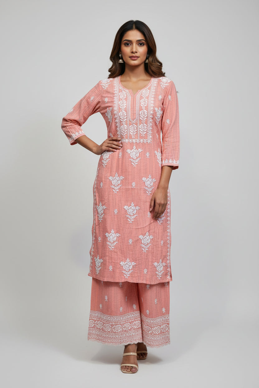 A woman standing on a sidewalk with wearing a peach pink colour traditional Lucknowi chikankari kurti with floral patterns and palazzo pants, holding a mobile phone and a bag.
