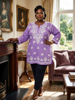 A woman stands in a classic indoor room wearing a lavender purple tunic top with white floral embroidery, paired with dark trousers and brown boots.