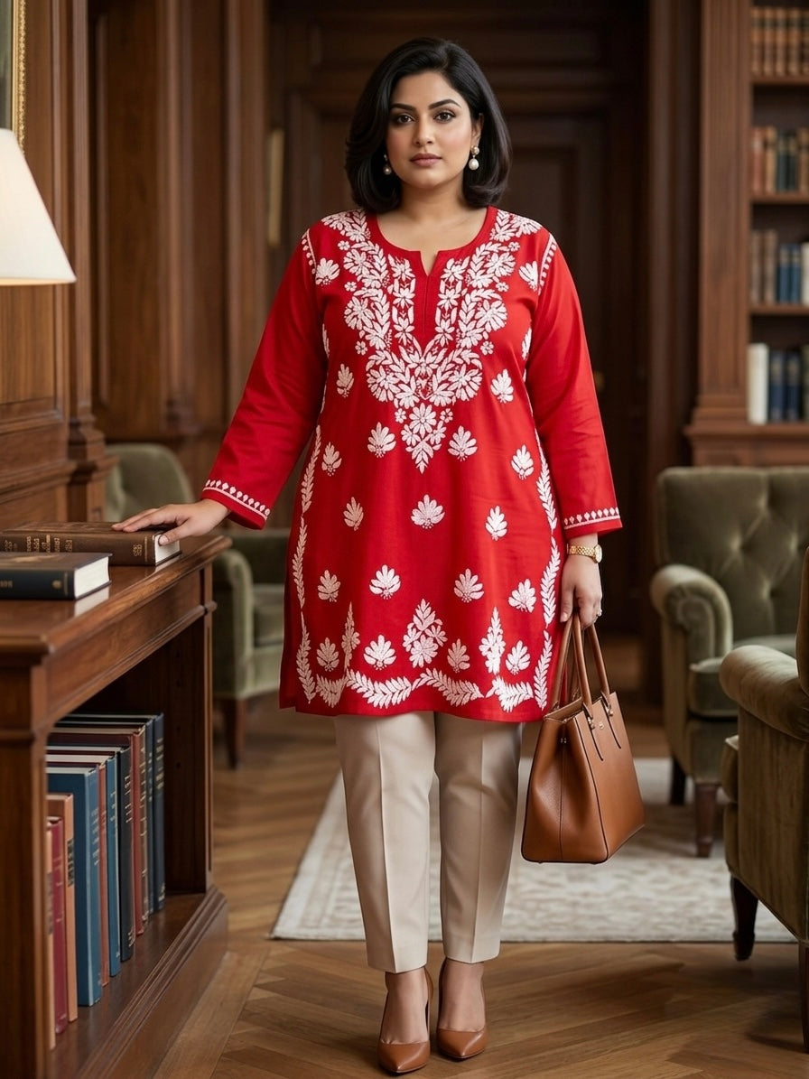 A woman stands in a library wearing a red kurta with white floral embroidery, paired with beige trousers and holding a brown leather handbag.