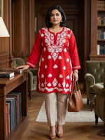 A woman stands in a library wearing a red kurta with white floral embroidery, paired with beige trousers and holding a brown leather handbag.