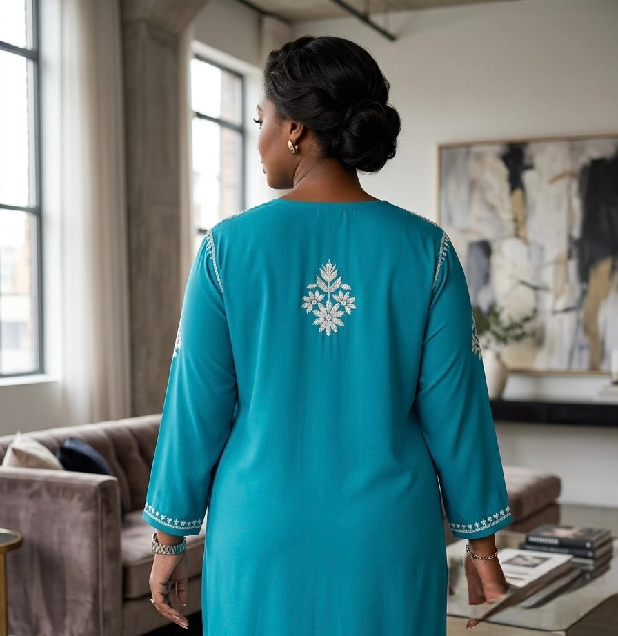 A woman viewed from behind wearing a turquoise blue tunic with white embroidered details on the back and sleeve cuffs, standing in a softly lit living room.