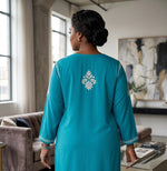 A woman viewed from behind wearing a turquoise blue tunic with white embroidered details on the back and sleeve cuffs, standing in a softly lit living room.