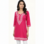 A woman modeling a bright pink kurta with white geometric embroidery around the neckline, sleeve cuffs, and hem, paired with white pants against a plain white background.