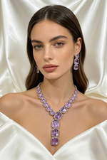 A woman wearing a purple gemstone necklace and matching drop earrings, posed against a draped white satin fabric background.