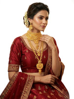 A woman wearing an ornate traditional Indian outfit in deep maroon with gold embroidery, styled with layered gold jewelry, a floral hair accessory, and earrings, set against a plain white background.