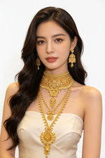 A woman with long dark wavy hair models an elaborate gold jewellery set, including a layered choker necklace, matching drop earrings, and a long pendant necklace, paired with a white strapless top in a jewellery store setting.