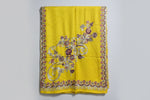 A yellow Kashmiri Aari embroidered marina scarf with tassels hanging at the ends, displayed on a wooden hanger with greenery in the background.