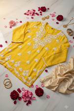 A yellow long-sleeve tunic top with white floral block prints, hanging on a wooden hanger against a light beige marble-textured background.