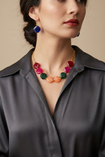 A young woman wearing the Facet Octagon Resin Necklace, showing off the balanced, mirrored arrangement of eight-sided beads in shades of peach, emerald, and ruby. She is also wearing the matching cobalt blue octagon-cut earrings, creating a clean, modern geometric jewelry look.
