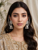 A woman wearing ornate silver-toned jhumka earrings with intricate beadwork and a matching embellished traditional outfit, shown in a close-up portrait against a soft cream background.