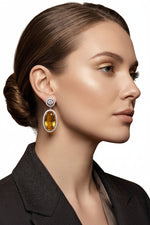 Luxurious oval yellow citrine drop earrings set in sterling silver with pave diamond halo accents, hanging against a soft, unfocused opera house-inspired gold velvet background.