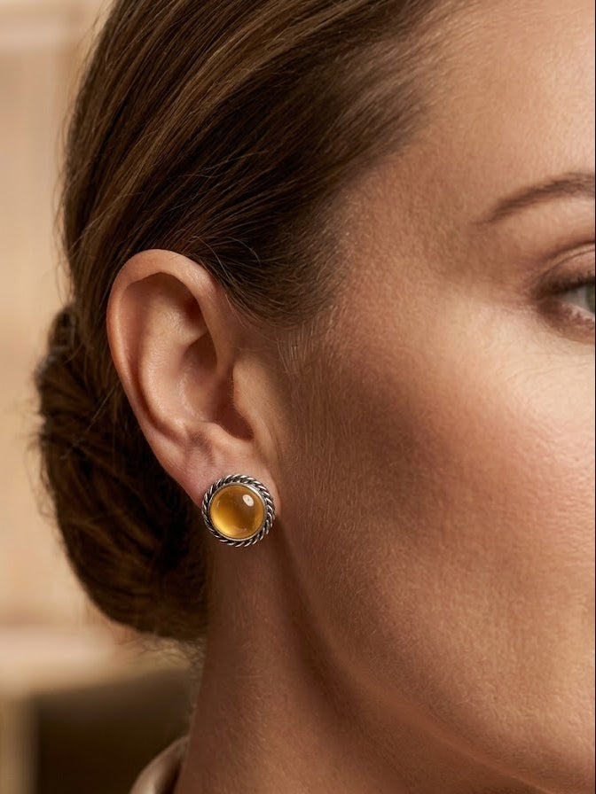 A close-up of a woman's ear wearing a small round stud earring with a textured silver-tone bezel and a warm amber-yellow cabochon center.