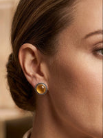 A close-up of a woman's ear wearing a small round stud earring with a textured silver-tone bezel and a warm amber-yellow cabochon center.