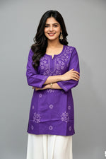 Front view of a woman wearing a royal purple cotton short kurti with white floral embroidery, a keyhole neckline, and a square hem line, paired with white trousers and standing with arms crossed.