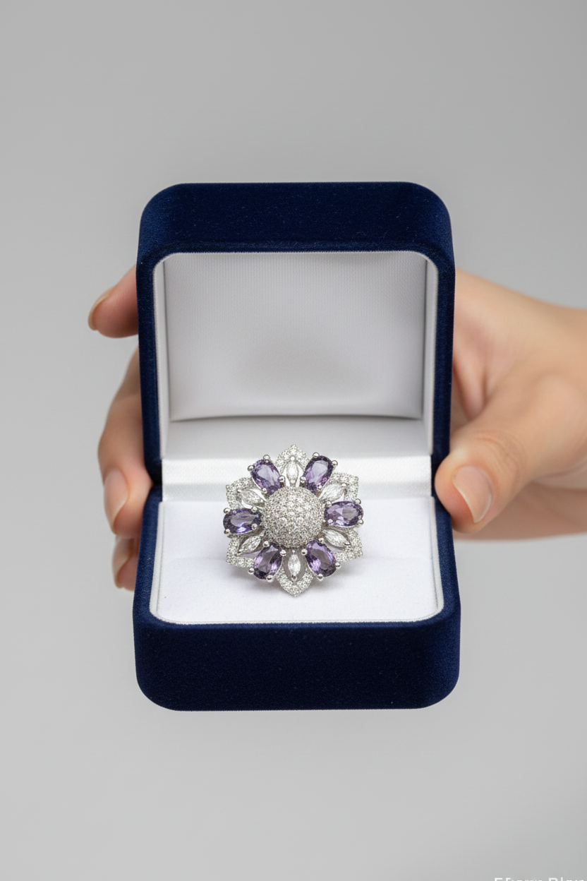 A silver-toned flower-shaped ring with purple gemstones and clear stones, presented in an open dark blue velvet ring box held by a hand against a neutral gray background.