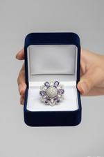 A silver-toned flower-shaped ring with purple gemstones and clear stones, presented in an open dark blue velvet ring box held by a hand against a neutral gray background.