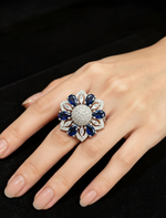 A close-up of a hand wearing an elaborate flower-shaped ring with dark blue oval stones and a silver-toned pavé center, set against a black background.