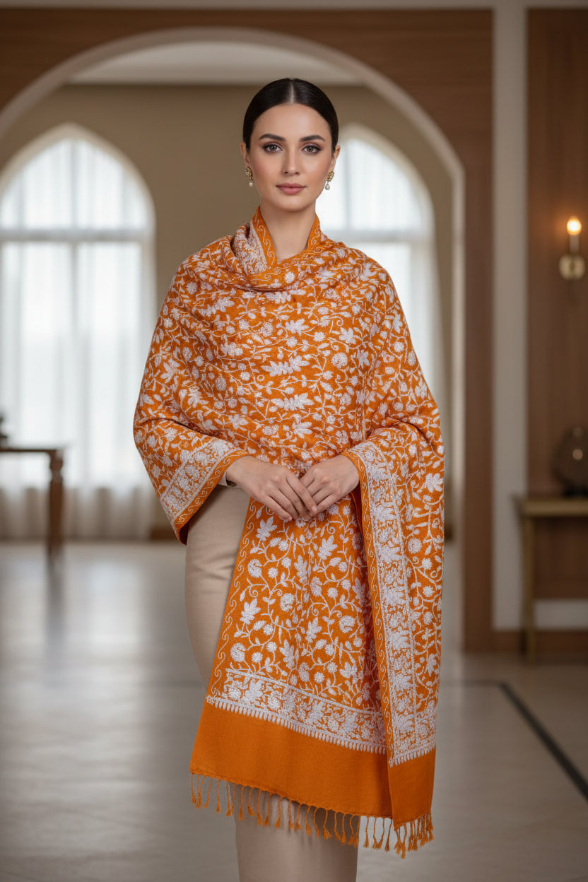 An orange Kashmiri pashmina scarf with white floral embroidery and tassels hanging at the ends, displayed on a golden background.