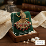 A gold-toned Indian-style bridal jewelry piece featuring a dancing woman motif and red stone embellishments, displayed on a green velvet jewelry box. The box sits on a wooden surface with pearls and books in the background.