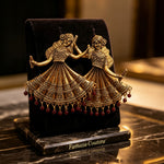 An ornate gold-toned jewelry display stand shaped like two dancing figures in traditional attire, holding hands and wearing layered embroidered dresses with dangling red bead drops, set on a black velvet cushion over a polished marble surface.