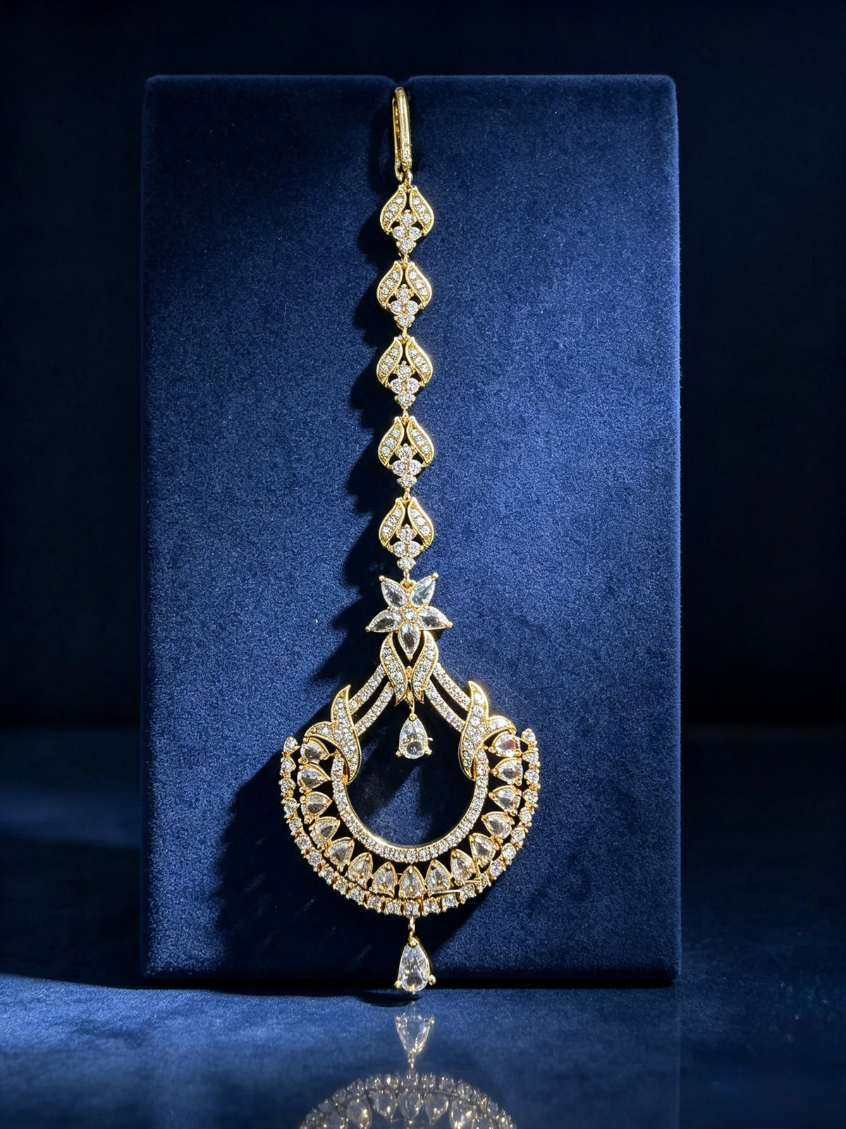 An ornate gold-toned traditional Indian jhumka earring embellished with numerous clear stones, displayed on a dark blue velvet jewelry box against a dark background.