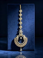 An ornate gold-toned traditional Indian jhumka earring embellished with numerous clear stones, displayed on a dark blue velvet jewelry box against a dark background.