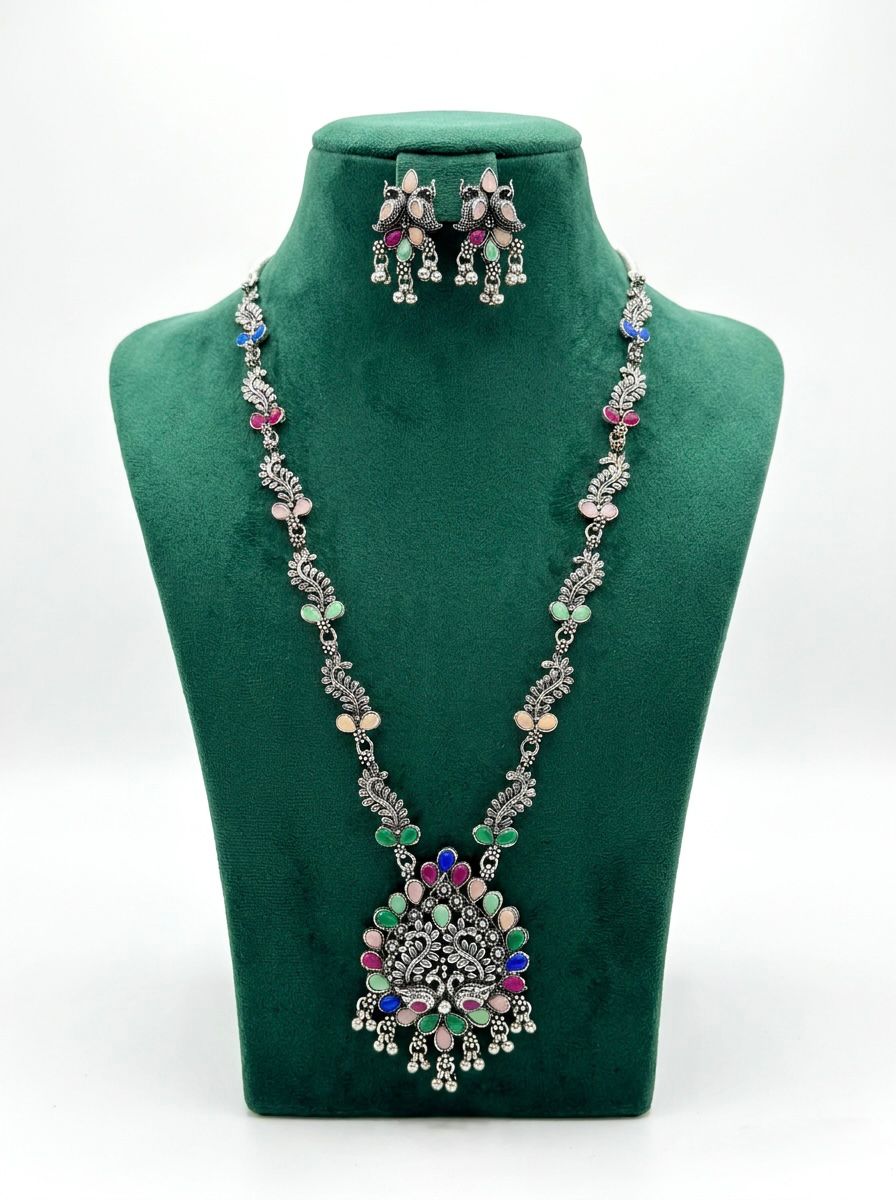 Antique oxidized silver long necklace and earrings set featuring an intricate peacock pendant and multicolor pastel stones on a green velvet display stand.