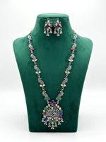 Antique oxidized silver long necklace and earrings set featuring an intricate peacock pendant and multicolor pastel stones on a green velvet display stand.