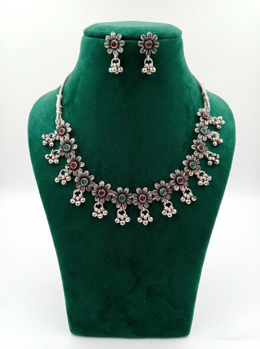 An antique silver-toned floral choker necklace and matching stud earrings set, featuring intricate lattice work, embedded ruby red and emerald green stones, and dangling cluster ghungroo beads, displayed on a green velvet stand against a pure white background.