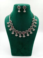 An antique silver-toned floral choker necklace and matching stud earrings set, featuring intricate lattice work, embedded ruby red and emerald green stones, and dangling cluster ghungroo beads, displayed on a green velvet stand against a pure white background.