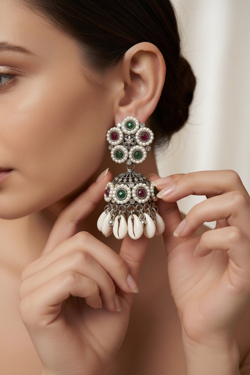 A close-up lifestyle shot of a woman wearing a large silver-toned dholakara-style earring adorned with green and red stones, while holding the earring with her fingers to show its details. The background is a softly blurred indoor setting.