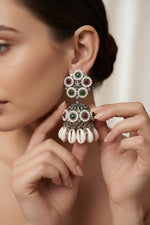A close-up lifestyle shot of a woman wearing a large silver-toned dholakara-style earring adorned with green and red stones, while holding the earring with her fingers to show its details. The background is a softly blurred indoor setting.