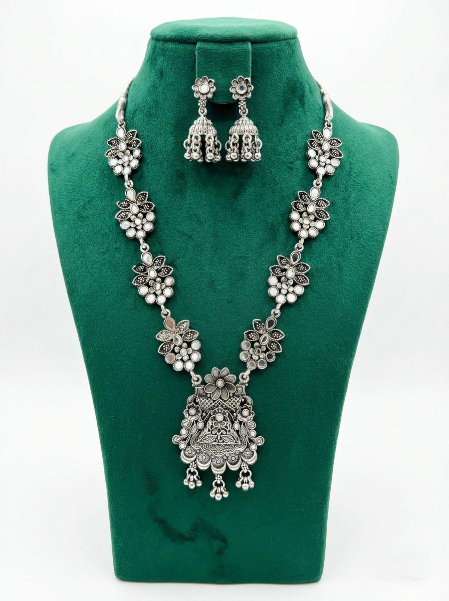 Antique oxidised silver necklace and matching jhumka earrings on a professional display stand, featuring intricate floral lattice mirror-work for luxury evening jewellery collections.