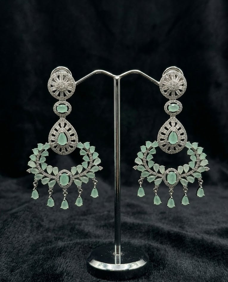 A sophisticated pair of Aqua Mist chandelier dangle earrings featuring rhodium-plated brass and intricate brilliant-cut stone detailing, showcased on a professional jewelry display stand against a textured black background.