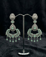 A sophisticated pair of Aqua Mist chandelier dangle earrings featuring rhodium-plated brass and intricate brilliant-cut stone detailing, showcased on a professional jewelry display stand against a textured black background.