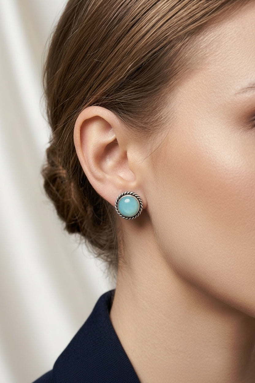 A close-up of a woman's ear wearing a silver-toned dangle earring with a round, light blue turquoise-like cabochon stone.