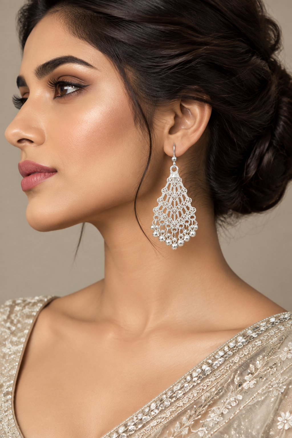 A close-up side profile of a woman wearing a large silver dangle earring with intricate floral lace detailing, paired with a silver embellished blouse against a neutral beige background.