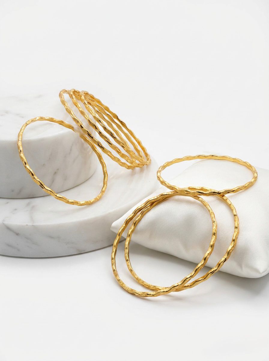 A professional product shot of eight micro gold-plated copper bangles with a textured wavy design. The bangles are elegantly arranged on a white marble pedestal and a soft white satin pillow against a clean white background.
