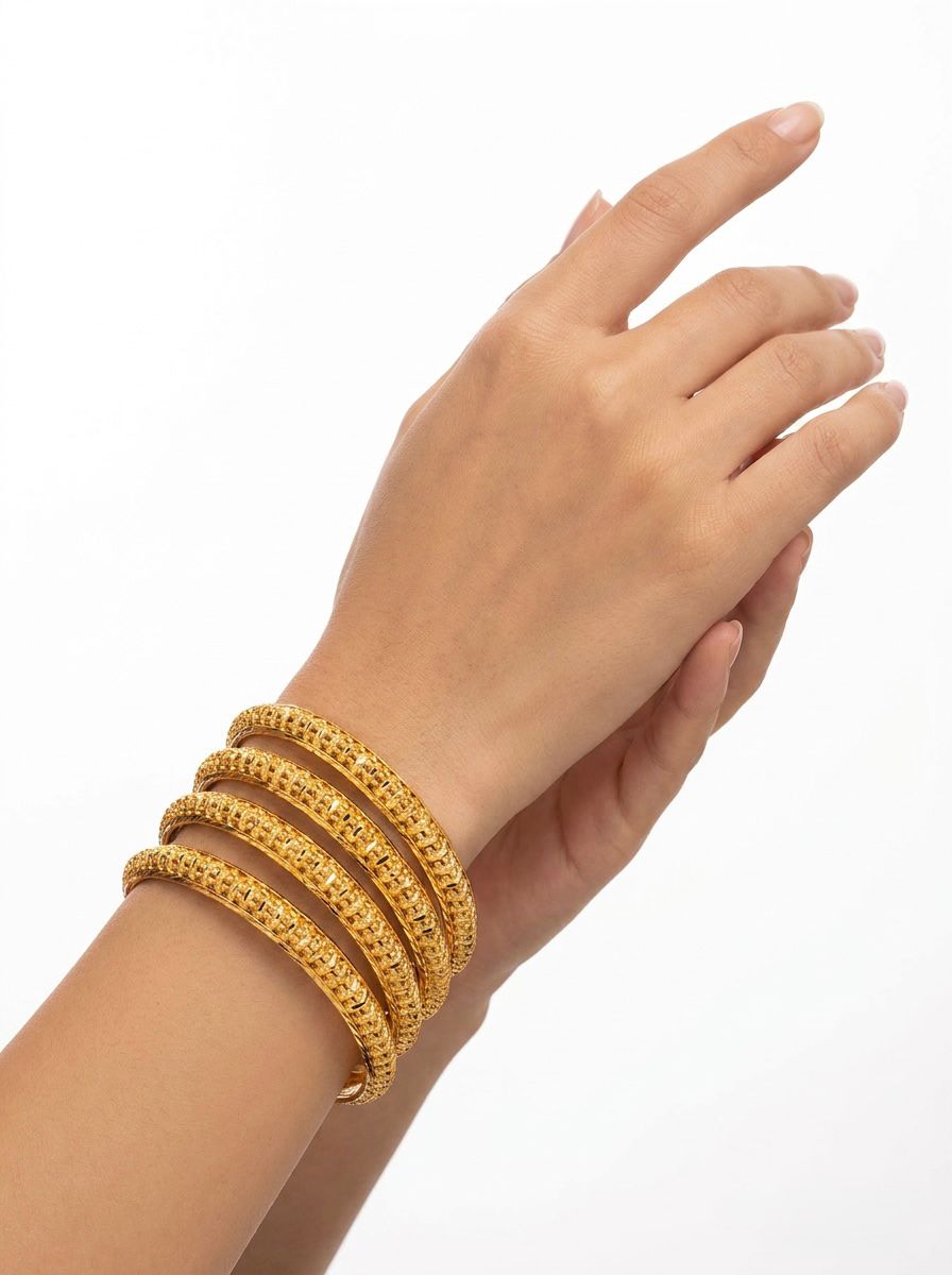 A bright lifestyle photograph showing a model's hands gracefully posed, wearing a set of four micro gold-plated copper bangles. The bangles feature a fine, repetitive beaded and lattice texture that catches the light, showcased against a clean white background.