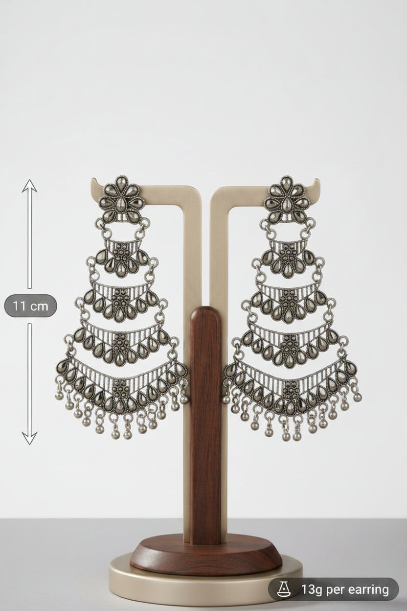 A pair of ornate silver-toned dangle earrings displayed on a wooden and metal stand, set against a dark brown draped fabric background with a blurred mirror in the distance.