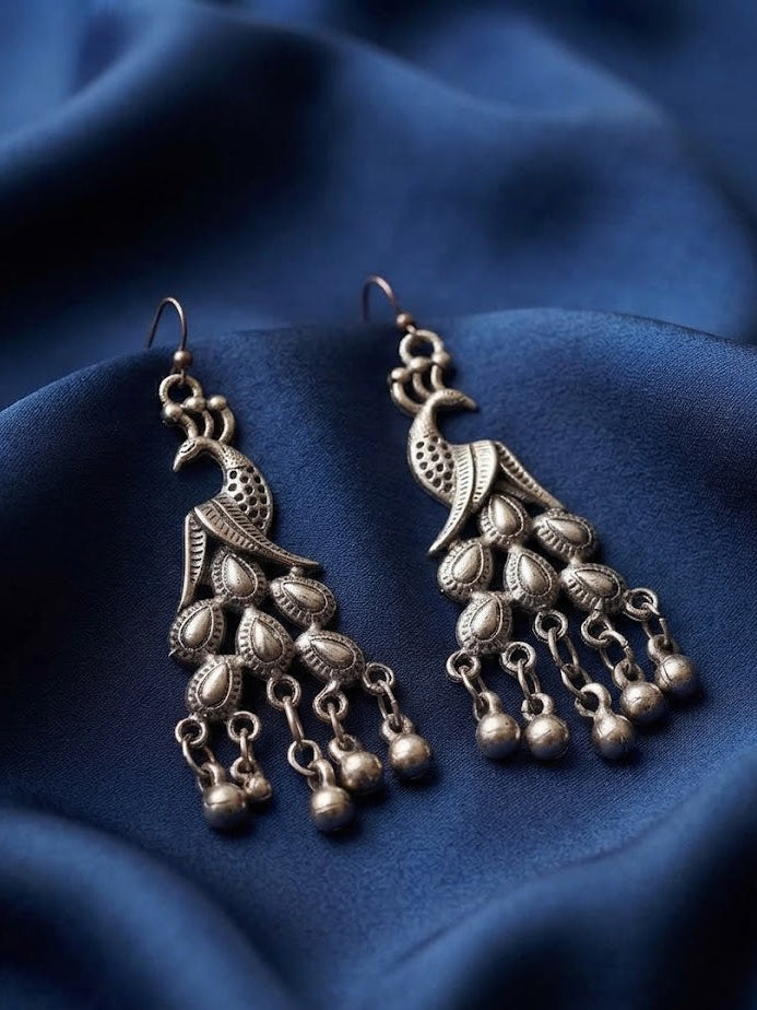 A pair of antique silver-toned peacock dangle earrings with hanging bell charms, displayed on draped dark blue fabric.