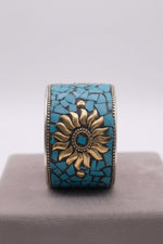 A vertical close-up shot of an artisanal, wide cuff bracelet featuring a detailed golden sunburst center surrounded by vibrant turquoise mosaic fragments, resting on a gray display block with a semi-transparent scale graphic indicating a width of 4.5 cm.