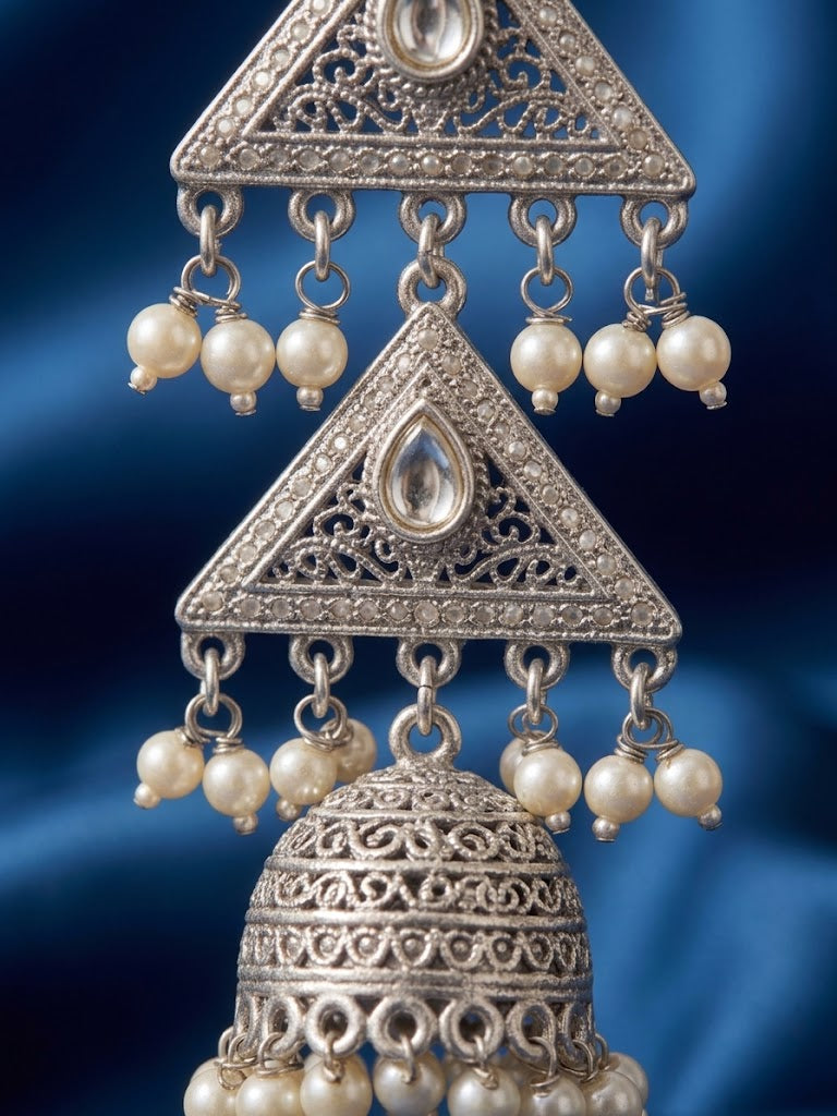 Silver-toned traditional Indian jhumka earrings with intricate filigree work, clear teardrop stones, and dangling cream-colored pearl beads, shown against a dark blue satin background.