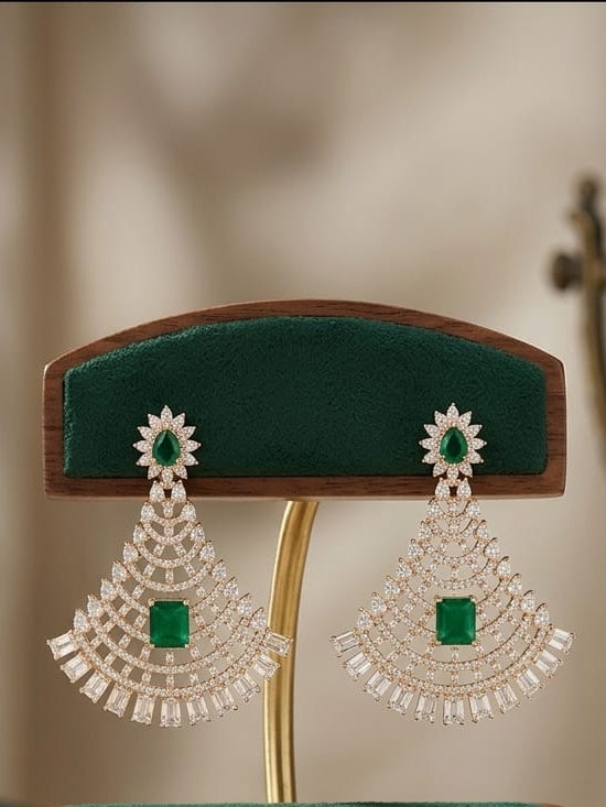A pair of large silver-toned statement earrings with green stone centers and clear crystal fringe, displayed on a wooden jewelry stand with green velvet cushions.