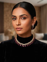 A woman with dark hair pulled back wears a gold-toned chain necklace with red teardrop stones and matching drop earrings, photographed indoors against a softly blurred neutral background.
