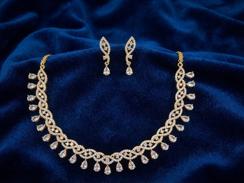 Gold-toned jewelry set featuring an ornate necklace and matching drop earrings, decorated with clear teardrop and marquise-shaped stones, displayed on a deep blue velvet fabric background.