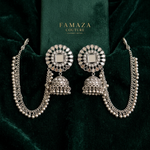 A pair of silver-toned Indian jhumka earrings with intricate circular patterns and dangling beaded chains, displayed on a dark green velvet surface with a green box labeled FAMAZA COUTURE in the background.