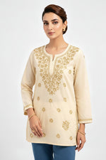 Front view of a model wearing a beige cotton short kurti featuring intricate gold embroidery on the yoke and scattered floral motifs, a keyhole neckline, and a square hem line, styled as a standalone short kurti without bottom wear.