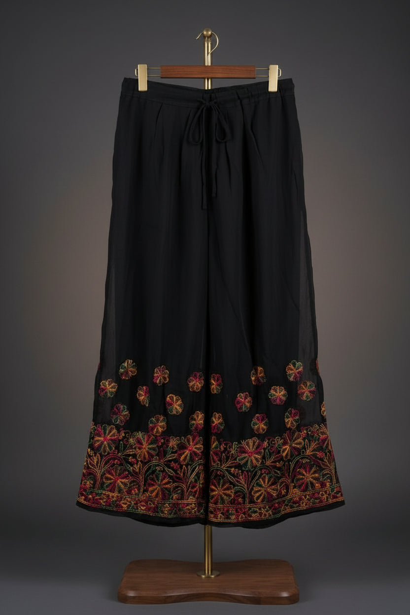 Front view of black wide-leg palazzo pants featuring vibrant multi-colored floral embroidery along the hem and an adjustable drawstring waistband.