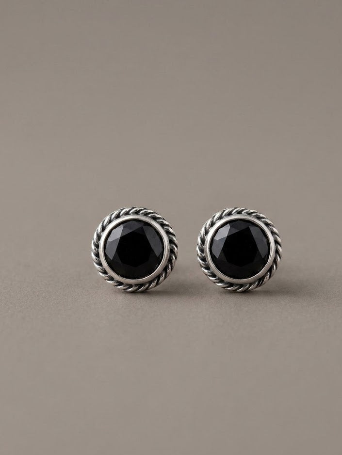 A pair of silver-toned round stud earrings with faceted black onyx centers, set in a textured rope-style bezel, on a neutral taupe background.
